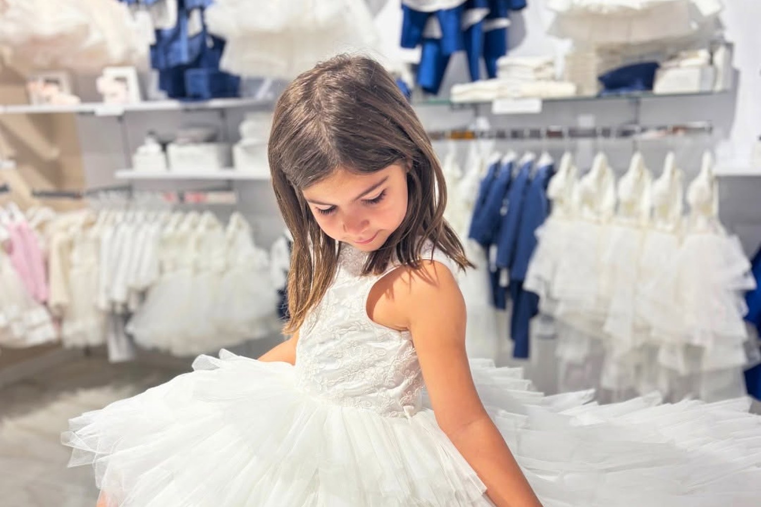 Bambina con abito bianco in tulle a balze e corpetto ricamato nel reparto cerimonia di MEY, tra abiti bianchi e blu