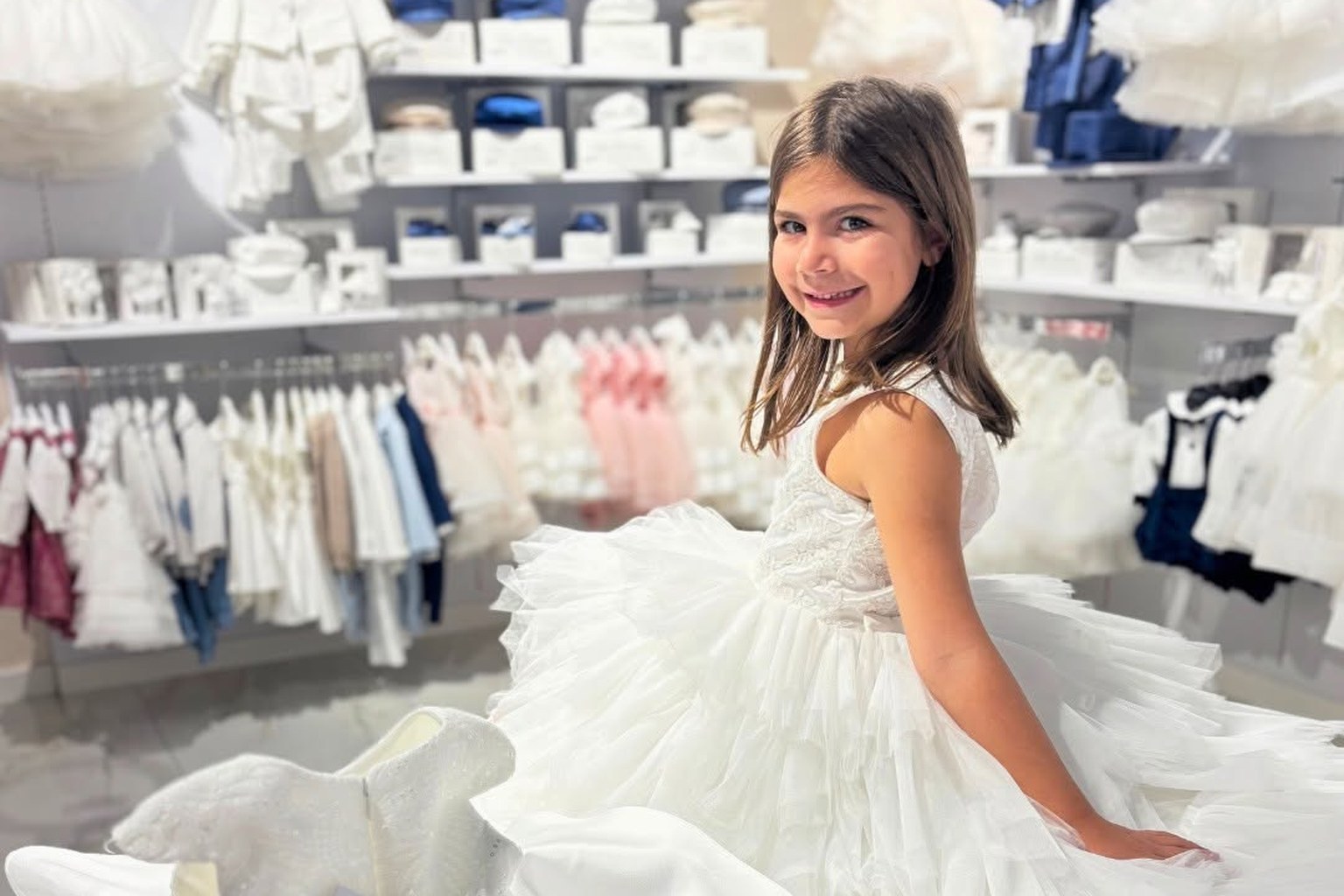 Bambina sorridente in abito bianco di tulle a balze nel negozio MEY, tra abitini da cerimonia appesi e scatole sugli scaffali